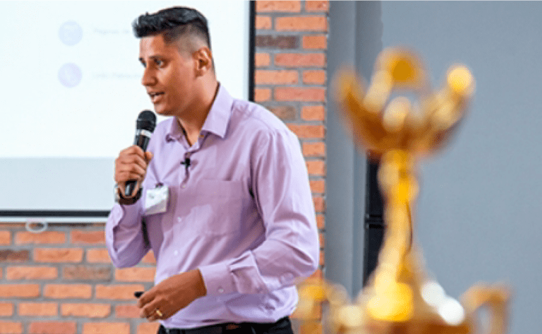 Rafael Coltro fala ao microfone durante uma apresentação, com um troféu em primeiro plano